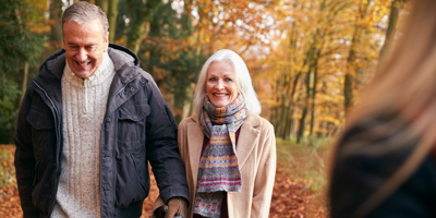 An older couple smiles while walking hand in hand along a wooded path in autumn, reflecting active aging, connection, and well-b